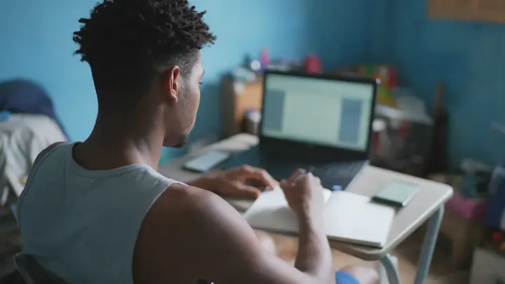 Jovem de costas numa escrivaninha de casa com notebook, celular e caderno, diante de uma parede azul