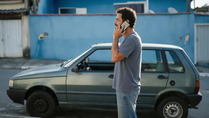 Homem ao lado do seu carro popular estacionado na rua, falando ao celular com expressão calma