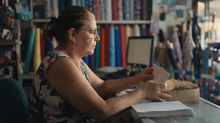 Dona de loja de tecidos conferindo fichas de clientes e um caderno no balcão, no fim do expediente