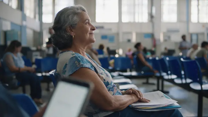 Senhora com uma pasta de documentos no colo aguardando atendimento em cadeiras azuis de um órgão público