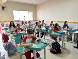 Aluno do Fundamental II escrevendo no caderno, com a turma ao fundo
