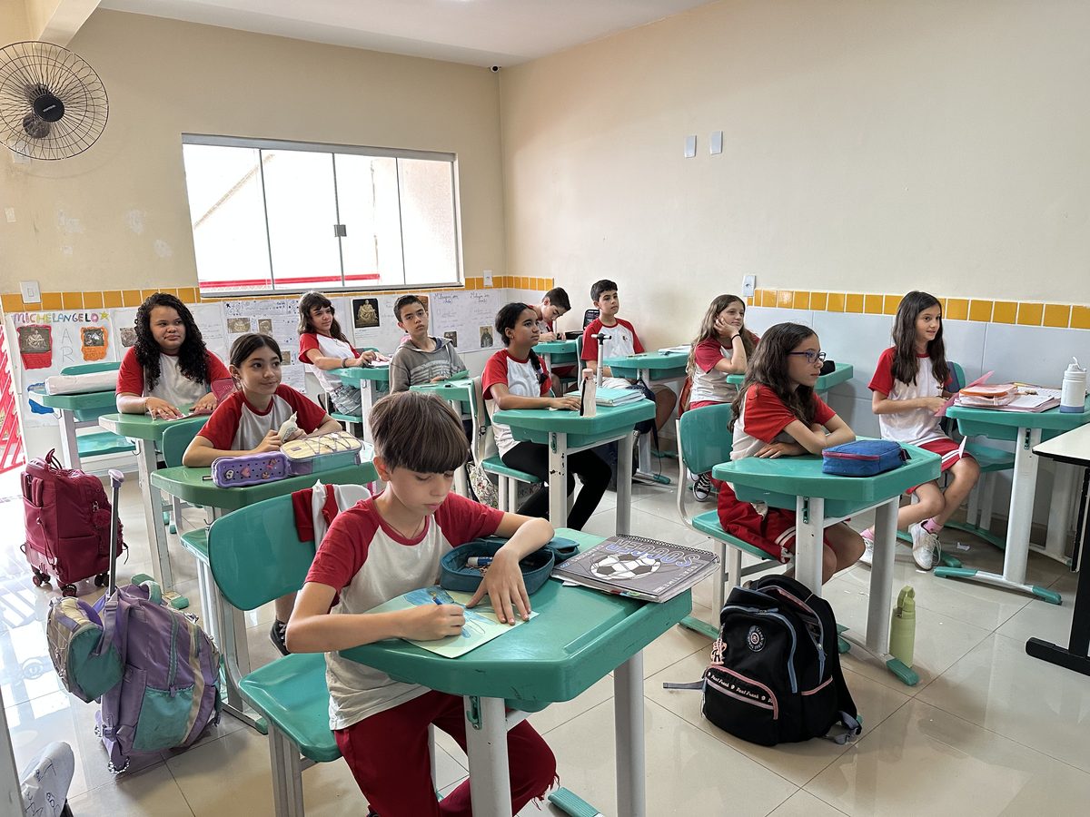Aluno do Fundamental II escrevendo no caderno, com a turma ao fundo